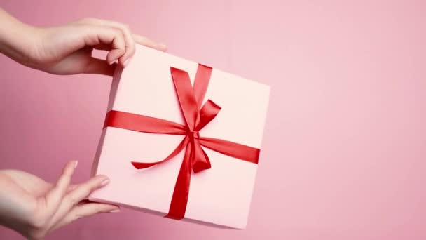 Kaukasische Hand zeigt Überraschung Geschenkbox mit roter Schleife Schleife isoliert auf rosa Pastell Wand - Filmmaterial, Video