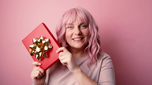 Portret szczęśliwej pozytywnej młodej kobiety w dobrym nastroju trzymającej pudełko prezentów odizolowane na różowym tle. Koncepcja świąt, uroczystości i kobiet - Materiał filmowy, wideo