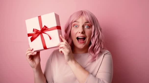 Celebración, feliz positiva emocionada mujer caucásica divertida sosteniendo presente caja de regalo con lazo de cinta roja posando aislado sobre fondo rosa. Estudio - Metraje, vídeo
