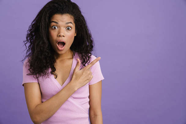 Молода чорна жінка висловлює несподіванку, вказуючи пальцем убік ізольовано на фіолетовому фоні
 - Фото, зображення