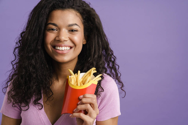 Young black woman smiling while eating french fries isolated over purple background - Фото, изображение