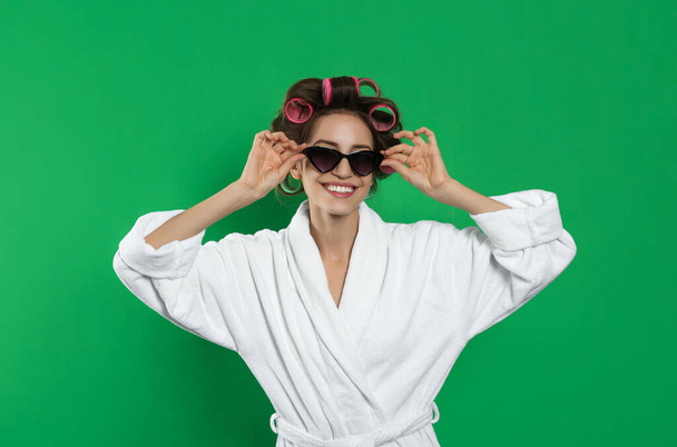 Beautiful young woman in bathrobe with hair curlers and sunglasses on green background - Foto, Imagem