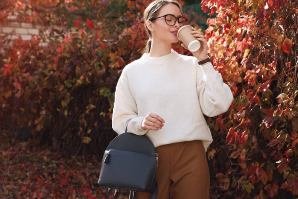 Beautiful young woman with stylish grey backpack drinking coffee in autumn park - Foto, Bild