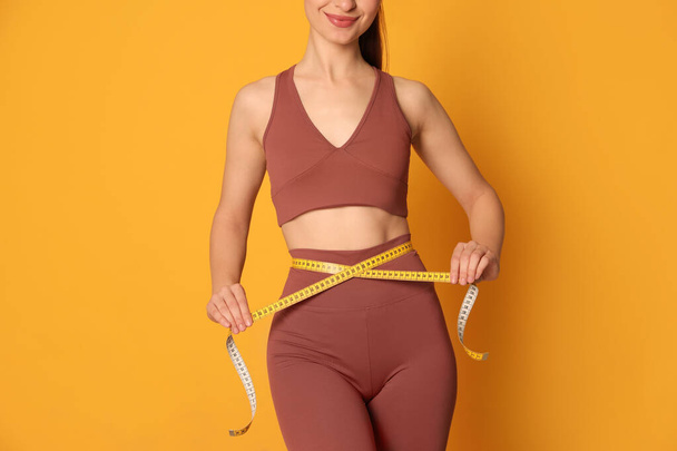 Happy woman in sportswear measuring waist with tape on yellow background, closeup - Zdjęcie, obraz