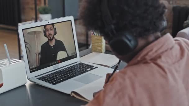 Au-dessus de l'épaule de Black Curly garçon d'âge élémentaire portant des écouteurs par-dessus l'oreille, assis au bureau à la maison dans l'après-midi, ayant des leçons en ligne avec le professeur asiatique masculin sur ordinateur portable - Séquence, vidéo