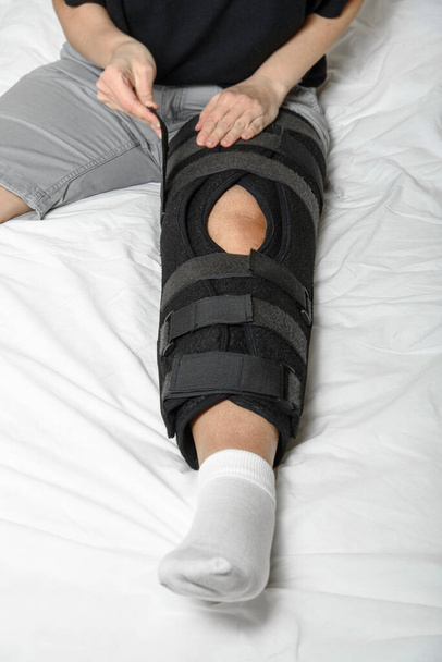 Mulher que fixa a órtese de velcro ajustável na perna quebrada sentada na cama. Usando cinta de perna após a lesão. Fecha a porta. Orientação vertical - Foto, Imagem