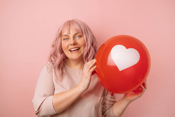 Belle jeune femme caucasienne des années 20 de bonne humeur souriante tenant coeur en forme de ballon rouge isolé sur fond rose. Fête de la Saint Valentin - Photo, image