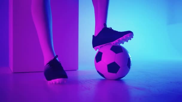 Primer plano del pie de un jugador de fútbol negro profesional de pie sobre la pelota en cámara lenta en la luz de neón azul-rojo del estudio. Jugador de fútbol brasileño pie en la pelota para posar - Metraje, vídeo