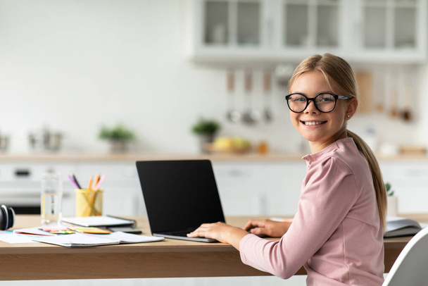 Cheerful caucasian teen schoolgirl in glasses look at camera at table with computer with blank screen - Photo, Image