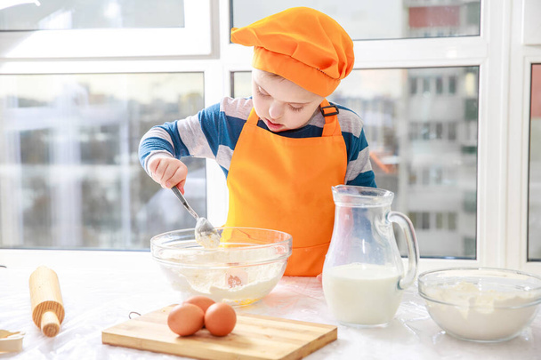 Um menino com síndrome de Down cozinha massa de farinha e leite na cozinha, trissonomia tipo 21, uma anomalia genética, uma criança vestida de chef.. - Foto, Imagem