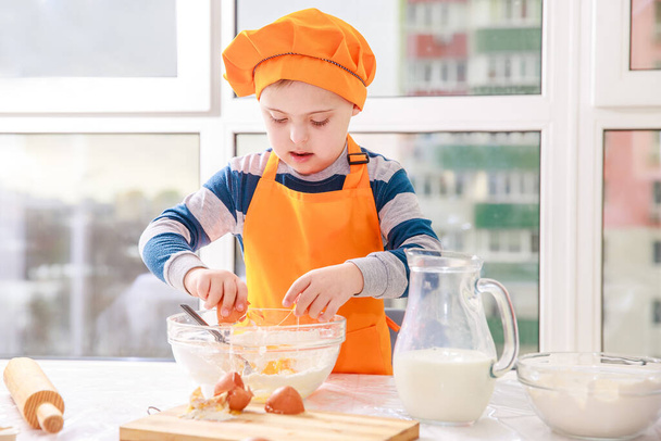 Un ragazzo con la sindrome di Down cucina la pasta di farina e latte in cucina, tipo 21 trisonomia, un'anomalia genetica, un bambino in costume da chef. - Foto, immagini