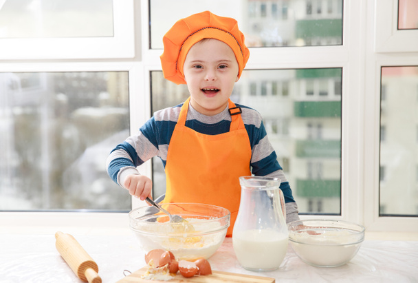 Um menino com síndrome de Down cozinha massa de farinha e leite na cozinha, trissonomia tipo 21, uma anomalia genética, uma criança vestida de chef.. - Foto, Imagem