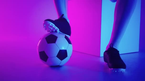 Primer plano del pie de un jugador de fútbol negro profesional de pie sobre la pelota en cámara lenta en la luz de neón azul-rojo del estudio. Jugador de fútbol brasileño pie en la pelota para posar - Metraje, vídeo