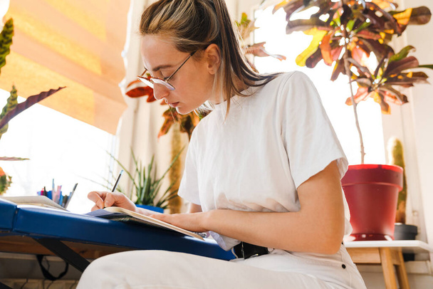 Young white woman in eyeglasses writing down notes while working indoors - Фото, изображение