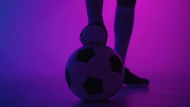 Stüdyonun mavi-kırmızı neon ışığı altında ağır çekimde topun üzerinde duran profesyonel bir siyahi futbolcunun ayağına yakın çekim. Brezilyalı futbolcu poz vermek için topa bastı - Video, Çekim