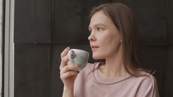 Chest up slowmo shot of young thoughtful Caucasian woman drinking coffee standing by window at home - Footage, Video
