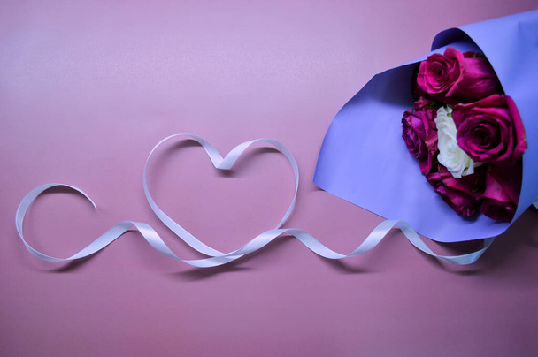Ein Strauß roter Rosen in lila Verpackung liegt auf rosa Hintergrund. Herz aus weißem Band - Foto, Bild