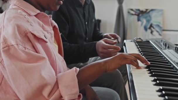 Side view low angle of cropped elementary-aged Black girl and young Asian man sitting on sofa in modern appartement overdag, leraar onderwijs student spelen synthesizer - Video