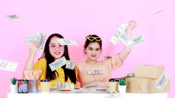 Retrato estudio toma de dos asiáticos feliz rico mujer rica startup pequeña empresa empresario socio de la mujer de negocios sentados juntos lanzando billetes de cien dólares lloviendo sobre fondo rosa. - Foto, imagen