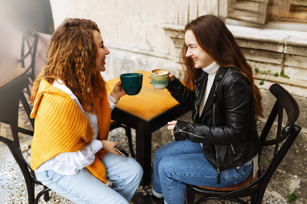 Fröhliche beste Freundin teilt Multimedia-Dateien auf Smartphones, verbringt Freizeit miteinander, lächelnde Hipster-Mädchen trinken Kaffee, sitzen im Terrassencafé - Foto, Bild