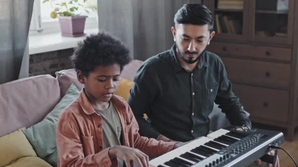 Μέσο αποφασισμένο μικρό μαύρο αγόρι σπουδάζει παίζοντας synthesizer με τον επαγγελματία δάσκαλο της Ασίας κάθεται στον καναπέ στο σπίτι το απόγευμα - Πλάνα, βίντεο