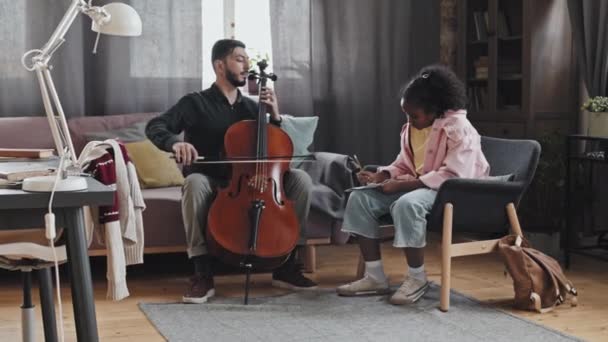 Full shot of Black curly-haired girl taking notes, watching professional male Asian musician playing double bass, sitting on couch at home - Footage, Video