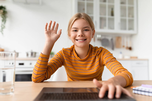 Веселая школьница-подросток смотрит на ноутбук, машущий рукой за столом в кухонном интерьере - Фото, изображение