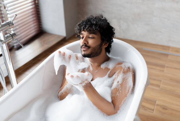 Handsome indian man blowing soap bubbles while relaxing in bathtub with foam, free space - Фото, изображение