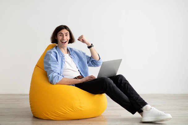Excited freelance man sitting in beanbag chair with laptop, celebrating online win, making YES gesture, free space - Photo, Image