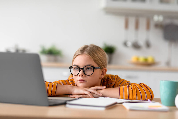 Tired serious pretty european teenage girl pupil in glasses looks at laptop, sits at table - Photo, Image