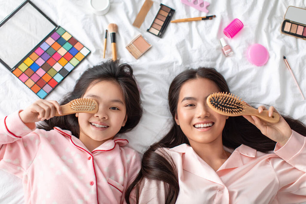 Boldog elégedett fiatal ázsiai nő és tizenéves lány, szoros szem fésűvel, feküdjön az ágyon kozmetikumokkal a hálószobában - Fotó, kép
