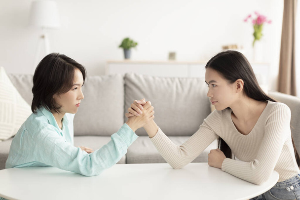 Mature Asian mother and adult daughter arm wrestling, looking each other in eyes, fighting at home - Foto, Imagem