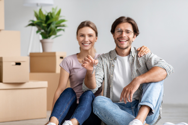 Satisfied caucasian young man in glasses and lady sit on floor show keys among cardboard box with stuff in room - Foto, immagini