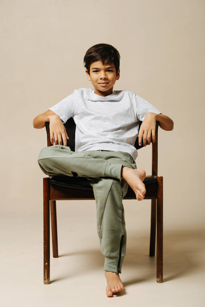 Cool confident indian boy sitting on an armchair in a room over beige background. He is leaning back, looking at the camera. - Фото, изображение
