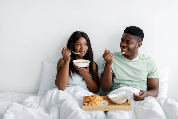 Cheerful millennial african american female and male wake up after night, eat porridge, enjoy good morning on bed - Photo, Image