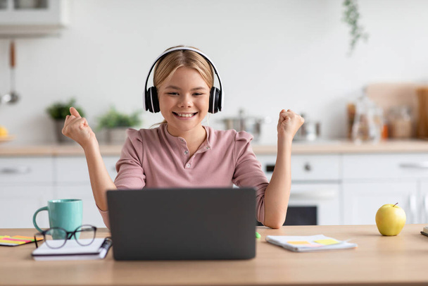 Happy glad caucasian teen girl student in headphones rejoices at successful result studying with laptop at table - Photo, Image