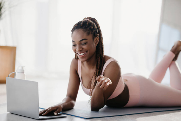 Happy African American woman in sports outfit lying on fitness mat, greeting personal trainer online on laptop at home - Fotó, kép