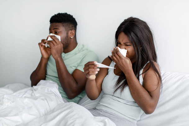 Λυπημένοι νεαροί αφροαμερικανοί σύζυγοι φυσούν τη μύτη τους σε χαρτοπετσέτες και κοιτάζουν το θερμόμετρο στο κρεβάτι στην κρεβατοκάμαρα - Φωτογραφία, εικόνα