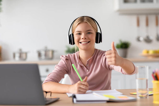 Smiling teen caucasian girl pupil in headphones show thumb up studying with laptop at table - Photo, Image