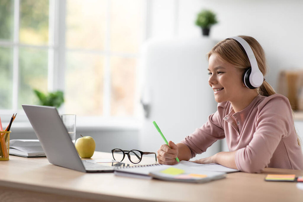 Iloinen eurooppalainen nuori tyttö oppilas kuulokkeet opiskelu kannettava tietokone pöydässä keittiössä sisustus, profiili - Valokuva, kuva
