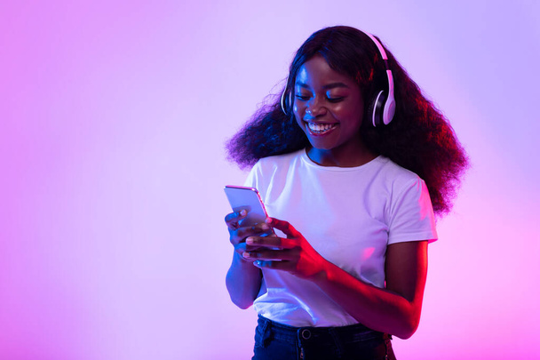 Alegre hembra negra milenaria en auriculares inalámbricos usando teléfono inteligente en luz de neón, espacio de copia - Foto, imagen