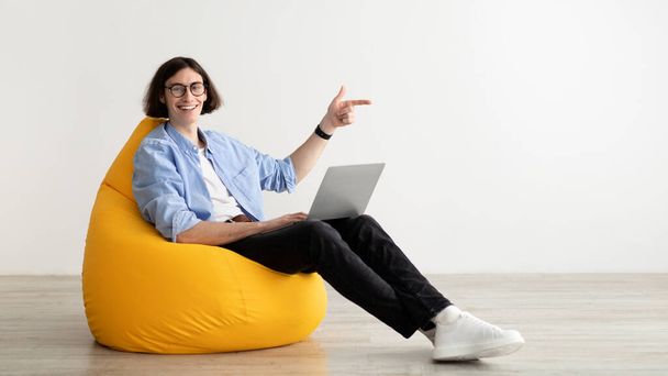 Excited freelance guy pointing fingers aside at copy space, sitting in beanbag chair and smiling at camera, panorama - Фото, изображение
