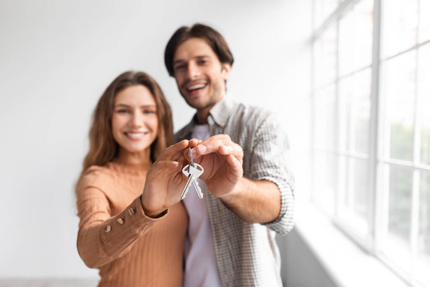 Счастливые кавказский мужчина и женщина около окна показывают ключи в пустой комнате, избирательный фокус - Фото, изображение