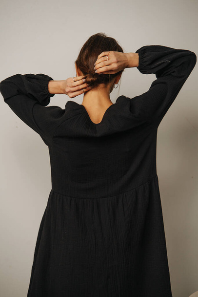 Backside of a woman in a black dress putting her hair in a knot, fixing it. Inside a room - Фото, изображение