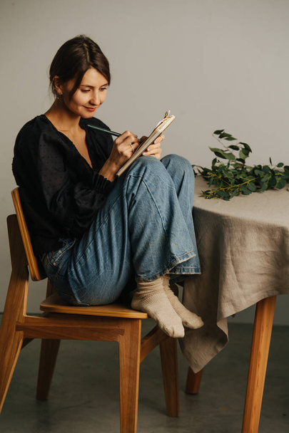 Happy woman sitting with legs up on a chair next to a round table, writing in a notepad. She's wearing mom jeans over black dress. Inside a room - Foto, imagen