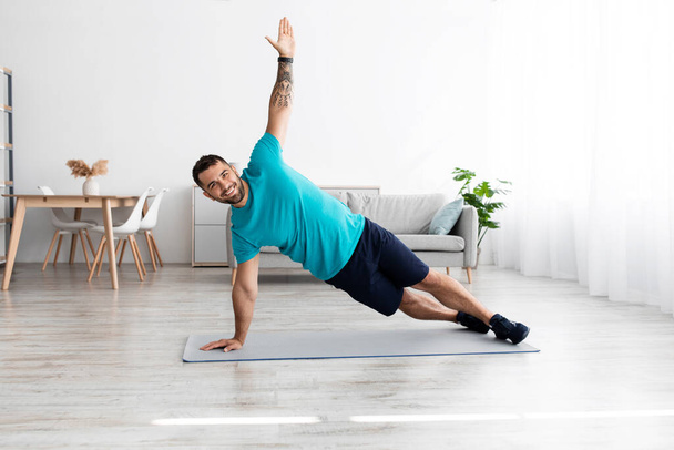 Smiling cheerful millennial european male athlete doing side plank at mat in minimalist living room - Photo, image