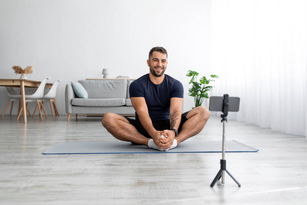 Happy european millennial guy sitting on mat looking at smartphone webcam recording sports blog in living room - Foto, afbeelding