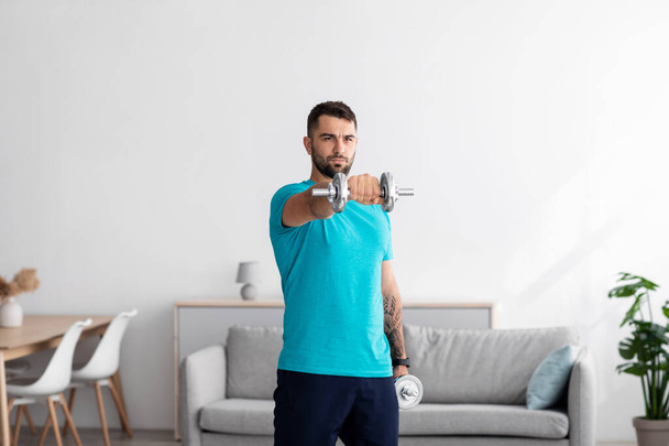 Serious millennial european man athlete in blue t-shirt raises dumbbell in minimalist living room interior - 写真・画像