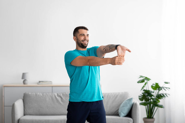 Sorrindo feliz millennial europeu cara em azul t-shirt fazendo exercícios e alongamento para os braços - Foto, Imagem