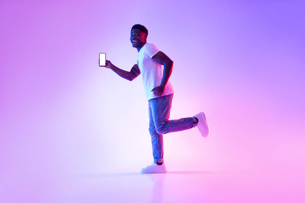 Jovem negro animado com smartphone rodando em luz de néon, mostrando mockup para design na tela do dispositivo móvel - Foto, Imagem